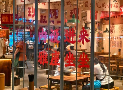 这是一张从室外拍摄的北京烤鸭店内部场景图，玻璃门上贴着醒目的红色手写风格标语“内设雅座 进来坐坐”，用接地气的话术招揽顾客。店内装修充满市井烟火气，摆放着木质桌椅，不少顾客正在用餐，还有工作人员在店内忙碌，墙上的标识和文字显示这是一家主打现烤北京烤鸭的餐馆，整体氛围热闹温馨，给人亲切的就餐氛围感。图中没有双关、谐音梗或暗示内容。