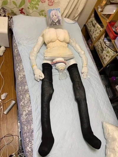 卧室场景中，床上躺着一个手工制作的人形玩偶。玩偶身体由枕头和布料拼接而成，穿着米色上衣和黑色过膝袜，双手戴着白色塑料手套。头部位置放置了一张动漫卡片，卡片上是一位粉色头发、穿着校服的微笑少女。背景可见 floral 图案窗帘、木质书架及收纳篮，地面散落着电线和小物件。整个作品展现了创意手工与动漫元素的结合。