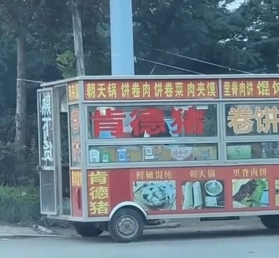 这是一张实拍的路边移动小吃餐车照片，餐车店主模仿知名快餐品牌肯德基的名称，将自己的餐车命名为“肯德猪”，红色大字“肯德猪”醒目展示在餐车正面，侧面也竖排标注了同样的店名；餐车多处标注了售卖的餐食品类，包括朝天锅、饼卷肉、饼卷菜、肉夹馍、里脊肉饼、馄饨等；餐车左侧玻璃的竖排文字写在玻璃内侧，从外部看呈现反向，实际内容为“概不赊欠”；餐车停靠在路边，背景是路边的树木。