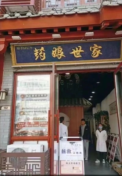 画面展示了一家具有浓郁传统中式建筑风格的店铺门面。红色飞檐搭配灰色瓦片，门楣上方悬挂着蓝底金字的招牌，左侧竖写'始于1852年'，中央为'药娘世家'四个大字，彰显百年老店的历史底蕴。店门敞开，门口站立几位身着白大褂的人员，门前摆放着一块写有'拯救上班族'的宣传牌，店内隐约可见传统中式装潢。整体场景充满古朴与现代结合的气息。