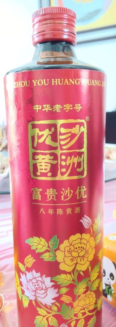 图片展示了一瓶红色的黄酒，瓶身主体为红色，配以金色文字和花纹。瓶身上方印有“中华老字号”字样，中央是“沙洲优黄”的方形标志，下方标注“富贵沙优 八年陈黄酒”。瓶身装饰有黄色和粉色的牡丹图案，背景隐约可见餐桌和部分食物，整体呈现出传统酒类商品的包装风格。