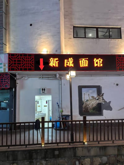夜晚的街边餐馆场景，上方是亮着红色和黄色霓虹灯的招牌，写着“新成面馆”，招牌左侧有一个醒目的红色箭头向下指向。箭头下方是卫生间入口，门口挂着“WC”标识。右侧墙壁上有一幅3D效果的长颈鹿壁画，长颈鹿的头部从画框中探出，形成有趣的视觉错位效果。整体画面展现了生活化的街边小店风貌，壁画设计增添了趣味性。