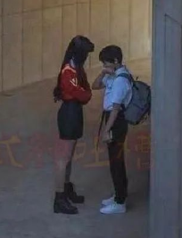这是一张利用视觉错位拍摄的趣味照片，画面中一位身着红黑套装的女生和背着双肩包的男生相对站立，由于拍摄角度的巧妙设计，男生站在靠近镜头的位置，女生站在稍远处，视觉上形成了女生远高于男生的夸张身高差，营造出搞笑有趣的反差效果。