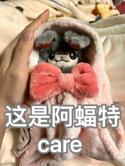 一只名为"阿蝠特"的可爱毛绒玩具被包裹在粉色的毯子里，上面系着一个大大的粉色蝴蝶结。玩具有着灰色的耳朵（内侧为红色）、黑色的眼睛和白色的面部，表情略带微笑。一只手轻轻托着这个包裹好的毛绒玩具，背景中可以看到床上的其他物品。整体画面呈现出温馨治愈的氛围。