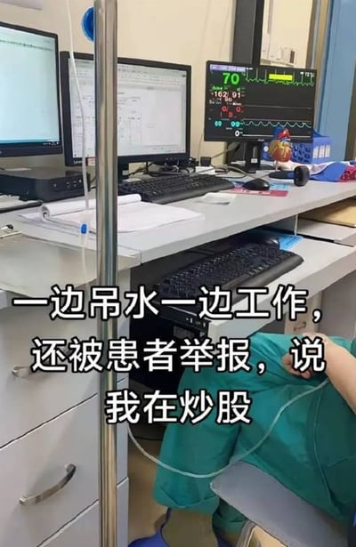 医院工作站场景：多张电脑显示器排列，其中一台显示心电监护仪数据（心率70，血压162/91）。画面下方可见穿着绿色手术服的医护人员坐在桌下，身旁有吊水支架。图中叠加文字：'一边吊水一边工作，还被患者举报，说我在炒股'。
场景通过幽默方式展现医护人员带病工作的辛劳，以及患者对医疗设备的误解（将工作用的监护仪屏幕误认为炒股软件），引发对职场压力与沟通错位的共鸣。