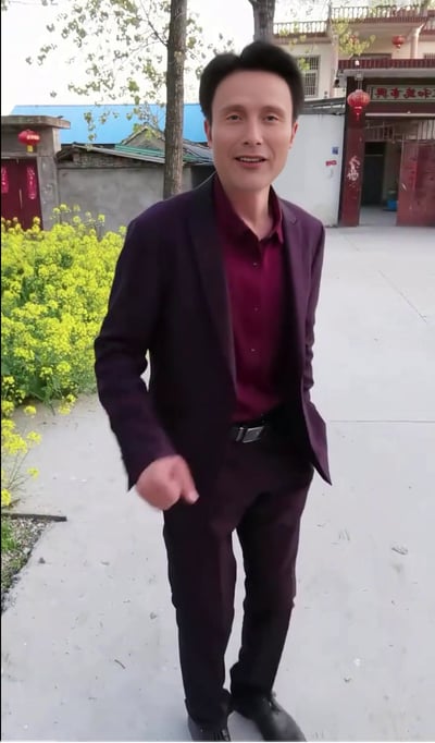 这是一张竖屏拍摄的户外实拍图，画面里一位身着深紫色西装套装、内搭酒红色翻领衫的中年男士站在乡村庭院的水泥路面上，他面带微笑，一只手插在裤袋，另一只手做出类似指向前方的动作。画面左侧是一片盛开的黄色油菜花，背景是挂有红色灯笼、带有中式门脸的乡村民居，整体氛围朴实又带着一丝正式感，展现出乡村生活里的日常瞬间。
