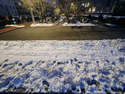 这是一张雪后夜晚的园区（大概率是大学校园）街景实拍图。前景是被踩踏过的积雪，雪地上写着一串由字母、箭头组成的趣味符号创作：“0→F(H(K(G))→H(K(G))→Horu(...)”，像是随性的文字游戏；背景中，道路两侧停满了电动车与自行车，树木根部积着未清扫的雪，路灯照亮了街道，路上有不少行人往来，还有车辆通行，整体氛围既有冬日雪后的清冷，又有夜晚园区的热闹烟火气。图中没有双关、谐音梗或暗示内容。