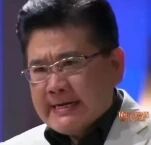 图片中是一位戴着眼镜的中年男子，面部表情显得非常激动，眉头紧锁，嘴巴微张，似乎正在表达强烈的情绪或说着什么。背景为模糊的蓝色调，整体画面像是从视频或电视节目中截取的片段。