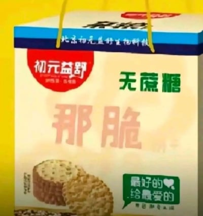这是一款仿冒知名品牌的山寨食品手提礼盒包装，顶部配有黄色提手。包装顶部边缘蓝底白字标注所属公司“北京初元益舒生物科技”，左上角红色标识内印有“初元益舒”字样，右上角用绿色字体标注“无蔗糖”，中间是醒目的浅粉色“那脆”字样，下方展示了两块带有谷物颗粒的饼干实物图，右下角绿色对话框样式的区域印有“最好的❤️给最爱的”宣传语，整体包装主打无蔗糖粗粮饼干的定位，试图通过仿冒知名品牌“初元”的名称来误导消费者，将其当作知名品牌产品购买。