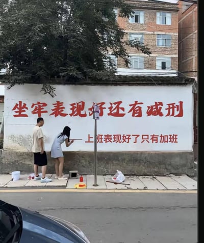 图片展示一面街头墙体，上面用红色大字写着“坐牢表现好还有减刑，上班表现好只有加班”。有两名年轻人在墙下描红补字，场景幽默又现实，讽刺了职场中的加班文化，把劳动与惩罚戏谑地类比在一起，带有明显自嘲和社会批判意味，适合用来表达打工人的无奈与调侃。双关梗：将“坐牢”表现好能减刑，与“上班”表现好只能加班并列，表达了现实生活的荒诞与反差。