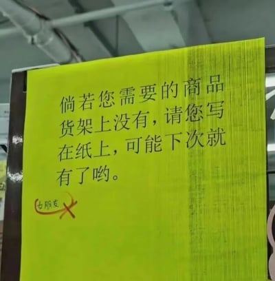 这是一张线下小超市或便利店的黄色提示牌照片，提示牌上的文字为店铺的补货提示：“倘若您需要的商品货架上没有，请您写在纸上，可能下次就有了哟。”，在提示牌的左下角，有顾客手写了“女朋友”的字样，并且被用红色笔划掉，形成了趣味的反差效果，用调侃的方式表达了“女朋友并非商品，无法通过补货获得”的现实，整体氛围轻松搞笑。