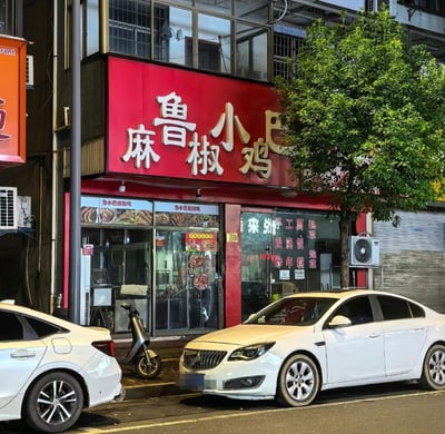 这是一张现实街景实拍图，画面主体是一家街边餐饮店铺，店铺红色门头的文字排版存在明显问题，原本的正确店名「鲁小巴麻椒鸡」因文字错位排列，容易被观看者错误断句读为谐音不雅的「麻鲁椒小鸡巴」，由此形成搞笑效果。店铺门口的人行道上停放着一辆电动踏板车，道路边还停有两辆白色轿车，店铺玻璃门上标注了该店经营的餐品种类，店铺旁还有一棵绿色的行道树。
