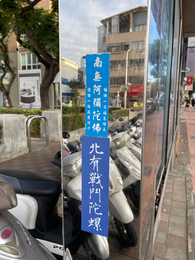 这张图片拍摄于城市街头，展示了贴在玻璃上的两张蓝色条幅标语。上方的标语是“南无阿弥陀佛”，下方则是“北有战吼花螺”，明显是一种恶搞式的对仗，利用宗教术语与网络流行语形成幽默效果。图中没有重叠内容或特殊场景，只是一种用文字制造的反差感。双关/谐音梗点：下部的“战吼花螺”用花螺这种贝类名称模仿了佛教念诵，略带戏谑神圣感；构造方式类似网络流行“xxx有xxx、yyy无yyy”式结构，具有地域和身份的幽默暗示。

整体风格轻松，表达了一种网络时代恶搞宗教语汇的常见现象，属于典型街头网络梗化标语。