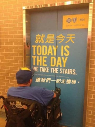 图片展示了一个充满讽刺感的场景：一位坐在轮椅上的人士停在医疗机构的电梯门前，而电梯门上贴着倡导人们走楼梯的标语，内容为“就是今天 TODAY IS THE DAY WE TAKE THE STAIRS. 讓我們一起走樓梯。”。这个标语旨在鼓励普通人多运动走楼梯，但完全忽略了行动不便的残障人士的需求，形成了强烈的反差，凸显了公共服务标语在考虑群体多样性上的疏漏，充满尴尬和讽刺意味。