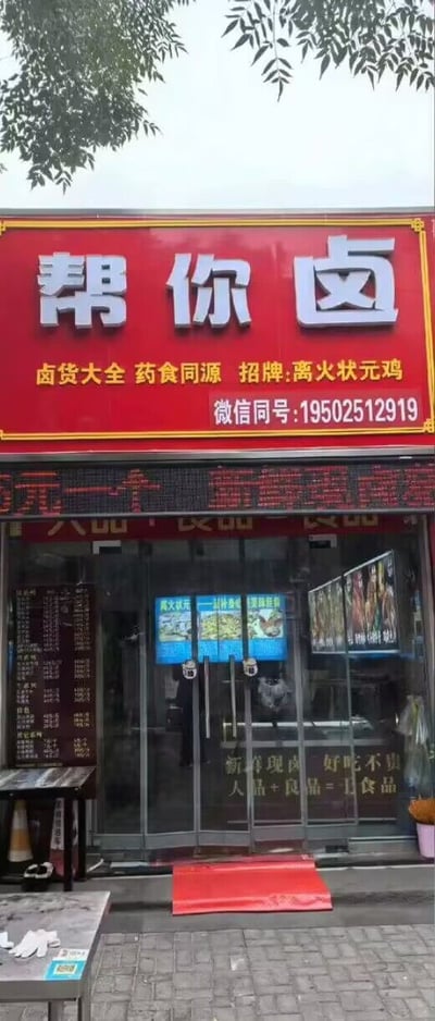 这是一家店铺的门头，最大的亮点是店名“帮你卤”，和网络热门梗“帮你囤”/“帮你盘”形成谐音，极具趣味性和年轻化气质。整张图以鲜艳红色底为背景，门牌醒目，还有“卤货大全”、“药食同源”、“招牌：离火状元鸡”等副标题，显示主营卤味食品，招牌菜名也有些夸张感。另外配有微信号，现代商业元素明显。

**双关&谐音梗说明**：店名“帮你卤”与“帮你囤”“帮你盘”等网络热词产生趣味联想，利用谐音和梗文化吸引年轻人眼球，是近年来线下门店常见的“梗式”营销套路之一。