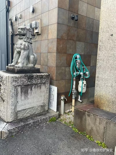 这是一处日本街头的建筑角落场景：左侧一尊日式狛犬（石狮子）石雕蹲坐于老旧石基座上，造型威严，是日本传统的守护石雕；旁边是贴有复古棕灰瓷砖的现代建筑墙面，墙面上挂着卷好的蓝绿色水管，还有水电表、插座等日常设施，基座旁立着一块日文使用说明牌，地面缝隙长出零星杂草，整体呈现出传统石雕与现代日常设施交融的平和生活化场景。
无谐音梗、双关或暗示内容。
