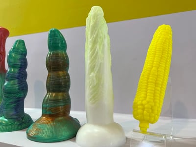 图片中展示了一组造型独特的彩色雕塑作品，整齐摆放在白色展示台上。左侧有两件绿色系雕塑，表面带有螺旋状纹理；中间是一件高耸的白色雕塑，形似白菜；右侧则是一个亮黄色的玉米造型雕塑。背景为黄色与灰色拼接的墙面，整体呈现出艺术展览中的静物陈列场景，色彩鲜明且造型富有创意。