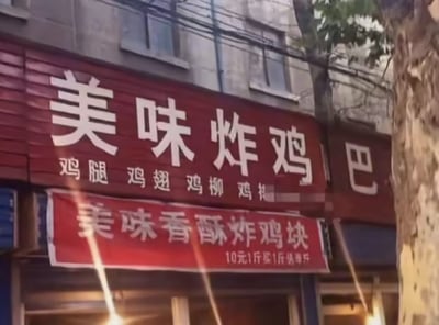 这是一张街边平民炸鸡店的实拍照片，店铺最上层是红色背景白色大字的门头招牌，原本完整文字应为“美味炸鸡吧”，但“吧”字的左半部分“口”被遮挡截断，仅露出右半部分“巴”，和前面的“炸鸡”连读会产生不雅谐音，形成笑点；大字下方标注了店铺售卖的品类，包括鸡腿、鸡翅、鸡柳，最后一个品类文字被遮挡；门头下方悬挂红色促销横幅，标注炸鸡块售价和优惠活动，画面右侧可见树干、老旧居民楼墙面和电线，整体是市井街边小吃店的场景，因招牌的意外截断产生的谐音梗在网络流传。