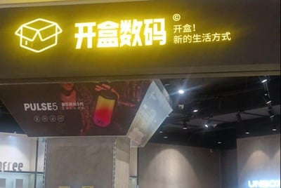 这是一张线下数码店铺的实拍图，画面上方是店铺的发光招牌：左侧是线条勾勒的打开的盒子图标，中间是醒目的“开盒数码”大字，右侧配有标语“开盒！新的生活方式”，整体灯光为暖黄色，视觉醒目；招牌下方悬挂着一款名为“PULSE5 音乐脉动5代”的音响宣传海报，海报上展示了这款带有渐变灯光效果的音响，凸显产品的影音属性；背景是店铺内部的灰色水泥质感墙面与嵌入式顶灯，整体风格简约现代，偏向新潮数码潮品店的定位，营造出时尚的购物氛围。