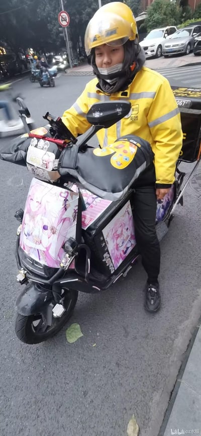 这是一张街头实拍照片，一位身着全套美团外卖黄色制服的外卖员坐在自己的电动车上。电动车的前部、侧面都贴满了粉色系的二次元美少女风格贴纸，车把的护手也装饰有可爱的卡通贴画，和专业的外卖工作装备形成了有趣的反差感，展现出这位外卖员在工作之余的二次元爱好，画面温馨又充满生活趣味。
无谐音梗、双关或暗示类内容。