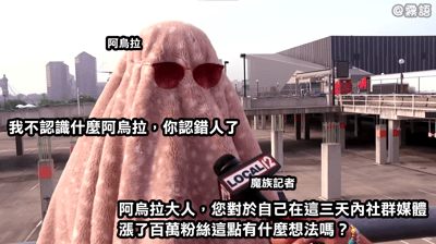 图片中一个头部被粉色毛绒物体覆盖、戴着红色墨镜的角色（阿乌拉）在停车场场景中接受采访。画面右侧有一名手持标有“LOCAL 12”字样麦克风的“魔族记者”向其提问，内容为“阿乌拉大人，您对于自己在这三天内社群媒体涨了百万粉丝这点有什么想法吗？”，而角色则回应“我不认识什么阿乌拉，你认错人了”。该图结合了动画《葬送的芙莉莲》中阿乌拉的“小提琴”梗，通过虚构的采访场景和角色否认身份的反差制造黑色幽默效果。