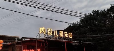 这是一张傍晚时分的户外实拍图，画面主体是一栋红砖结构的临街店铺，店铺屋顶的金属支架上安装着醒目的黄色发光字体招牌，内容为“内设儿童乐园”，旨在吸引带孩子的顾客。店铺外墙可见空调外机，下方还有部分可见的红色发光字样，左侧悬挂着中式灯笼。背景是阴沉的天空，多组交错的黑色电线横贯画面，右侧则是茂密的深色树木，整体场景充满市井休闲气息，推测该店铺为餐饮类场所，主打亲子友好的消费体验。