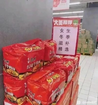 这是一张超市内的实拍静态图，画面主体是堆叠起来的多组康师傅红烧牛肉面五连包，旁边立着一块红色边框的推荐牌，上面写着“大美推荐 女生冬季滋补精选”。用原本属于养生滋补品的推荐话术来介绍速食泡面，形成强烈的反差感，制造出搞笑的效果，调侃当代年轻人冬季可能凑活饮食，将泡面戏称为“滋补品”的无奈又诙谐的生活状态。图片右下角带有“@meme图吧”的水印，表明该图在该网络平台传播。