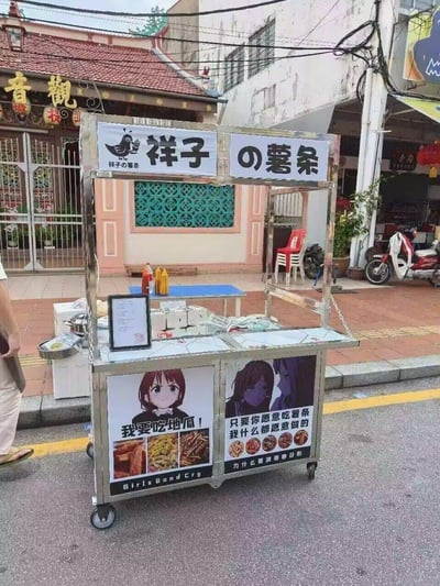 这是一个摆放在南洋风格街边的移动不锈钢薯条小吃摊，摊名定为「祥子の薯条」。摊位正面贴有两张二次元角色宣传海报：左侧是《GIRLS BAND CRY》的井芹仁菜，配文「我要吃地瓜！」，下方搭配地瓜类炸物的实拍图；右侧是《孤独摇滚！》的伊地知虹夏，配文「只要你愿意吃薯条 我什么都愿意做的」，下方展示了多种口味薯条的样式。摊位台面上摆放着调料瓶、电子菜单、包装材料等经营用品，背景是带有中式骑楼风格的建筑、停放的摩托与户外桌椅，整体场景充满生活气息，同时通过热门二次元番剧的角色梗做宣传，兼具网感与趣味性。