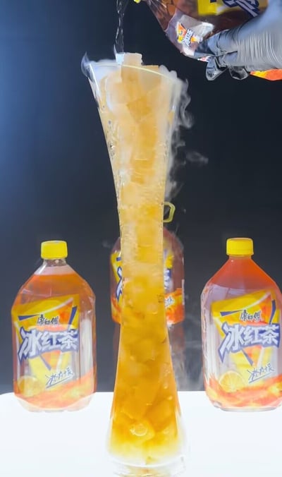画面主体为一个极高的窄口玻璃容器，容器内装满冰块，正有一只戴灰色手套的手将康师傅冰红茶倒入其中，容器边缘冒着白色冷气，视觉上凸显出饮品的冰爽感；容器两侧及后方还摆放了多瓶大瓶装康师傅冰红茶，整体画面突出了远超常规分量的冰红茶饮品，视觉冲击力较强。