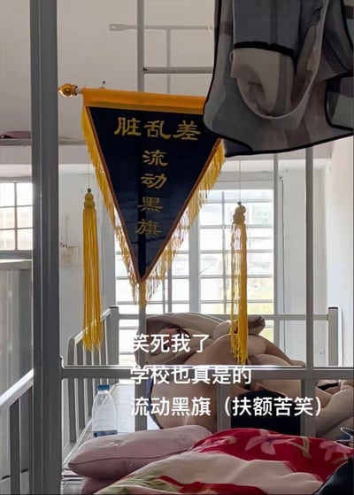 这是大学宿舍的真实场景记录，上下铺床位框架上悬挂着学校颁发的“脏乱差流动黑旗”，床铺区域被褥凌乱，图中配文“笑死我了 学校也真是的 流动黑旗（扶额苦笑）”，生动展现出学生面对这面反向激励的黑旗时，既觉得好笑又有些无奈的心情，用幽默的方式吐槽校园宿舍卫生评比的有趣操作。