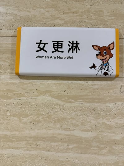 这是一块安装在米黄色大理石墙面上的恶搞标识牌，标识牌主体为白色，两侧带有黄色包边。标识牌上方是中文文字“女更淋”，下方配有英文翻译“Women Are More Wet”，该翻译属于完全错误的字面直译，将原本应该是“女更衣室”的错误文字进行了低俗化的错误翻译，产生了怪异的含义。标识牌右侧绘制了一个穿着白大褂、佩戴听诊器的卡通小鹿形象，小鹿面带笑容，左手叉腰，右手做出邀请的手势，整体风格诙谐搞笑，标识牌整体为恶搞性质，通过错误的文字和翻译制造荒诞的喜剧效果。