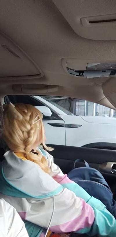 车内后排视角拍摄的场景，一位金发编成侧马尾的女孩坐在副驾驶座上，穿着蓝白粉渐变色调的连帽外套，正扭头望向车窗外的白色汽车。她的头发上别着银色发卡，身旁座位上放着一个深蓝色背包。车内可以看到车顶的阅读灯和遮阳板，窗外似乎是一个停车场或汽车展厅环境。