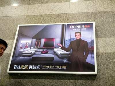 这是一张设置在地铁站水磨石墙面上的欧派家居商业广告，画面采用左右分景设计：右侧是品牌代言人胡歌，他身着深棕色西装搭配黑色高领内搭，神情沉稳自信，一只手插在裤兜，另一只手伸出，做出展示的动作；左侧呈现了一个现代风格的客厅场景，浅灰色的组合式沙发、深色矮凳、圆形红棕色茶几搭配简约地毯，背景是带有粉色氛围灯的电视墙，延伸出的空间里还能看到衣柜等家居，整体风格简约高级。广告上方标注“OPPEIN欧派 整家定制+家装”，底部配有宣传语“看过欧派 再装家 | 一体化设计 一揽子搞定”，同时附带品牌客服电话、官网以及股票信息，主打一站式的家装与整家定制服务。