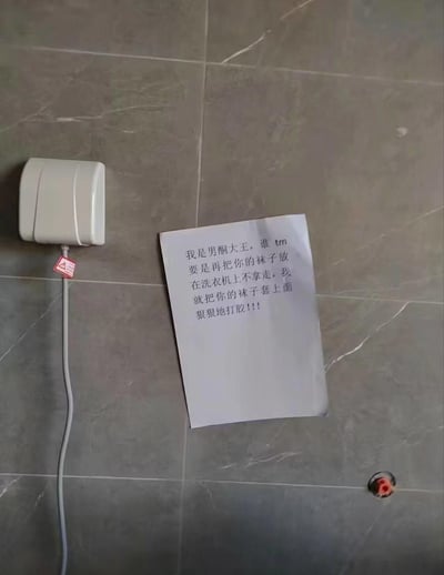 图片展示了一面灰色瓷砖墙，左侧安装着一个白色的电器设备（疑似烘干机或感应器），其下方垂着一根白色电源线并系有红色标签。设备右侧的墙面上贴着一张手写纸条，内容为：“我是男酮大王，谁 tm 要是再把你的袜子放在洗衣机上不拿走，我就把你的袜子套上面狠狠地打胶!!!”。整体场景像是家庭或合租公寓的洗衣区域，纸条内容以幽默夸张的语气表达了对他人将袜子遗留在洗衣机上的不满与警告。