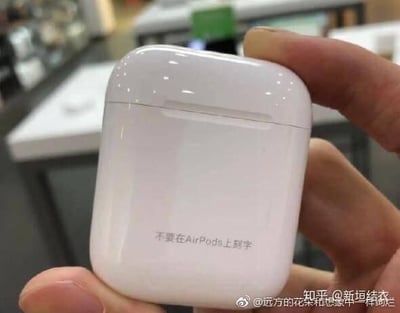 一张近距离拍摄的照片，展示了一只手握着白色的AirPods耳机盒。盒子表面用激光雕刻着一行中文文字：“不要在AirPods上刻字”，形成强烈的反讽效果——用刻字的方式警告不要刻字。背景是模糊的商店柜台，暗示可能拍摄于电子产品零售店。