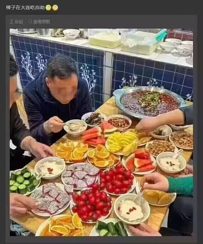 这是一张记录多人在大连享用自助美食的实拍图，画面中的餐桌上摆满了丰富餐品：有切分好的橙子、圣女果、西瓜、猕猴桃等新鲜水果，还有多种凉拌小菜、主食，以及一大锅红汤类餐品。数位食客围坐在桌前，有的正用勺子用餐，有的伸手取餐，氛围惬意。图中顶部配有文字“棒子在大连吃自助”，其中一位食客的脸部做了马赛克处理，背景是带有花纹的蓝白瓷砖墙面，还能看到一些餐具容器，整体呈现出熟人聚餐享用自助的日常场景。