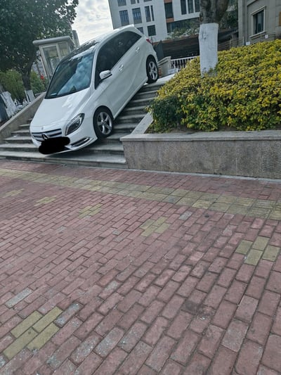 一张城市住宅小区内的实拍照片，画面中央是一辆白色奔驰轿车意外驶上了一段石质台阶。车辆前半部分卡在台阶上，车头朝向下方，车身倾斜，前轮悬空在台阶边缘，后轮可能仍在较高处的平台上。台阶旁有修剪整齐的绿色灌木和一棵树干涂白的树木，背景可见多层居民楼和小区门卫亭，地面为红黄相间的砖铺人行道。车辆前脸的车牌被黑色色块遮挡处理。整体场景呈现出一种意外事故后的荒诞感，推测可能是驾驶员操作失误导致车辆失控冲上台阶。