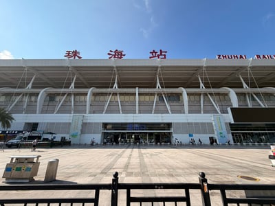 这是一张在晴朗白天拍摄的珠海站正面全景照片。画面采用对称式构图，站房占据中上部，前景是宽阔的石材铺装广场，最下方有一道黑色金属护栏。珠海站主体为现代风格，外立面以白色和灰色板材为主，顶部檐口上方立有醒目的红色“珠海站”三个大字，右侧露出英文“ZHUHAI RAIL”字样。屋顶由多组白色斜拉钢索和拱形钢结构支撑，线条富有张力。站房正中央是玻璃门入口，门楣上方有蓝色“进站口”三字；入口两侧墙柱上挂有竖幅彩色宣传海报，右侧墙面嵌有一块黑色电子显示屏。广场地面由浅灰、米黄色地砖铺成，阳光照射下阴影清晰，可见天气晴好。左下角放置一组不锈钢分类垃圾桶和一个方形金属柱，左侧远处停有一辆白色车辆，并可见棕榈树；进站口前有多名旅客拖着行李行走或进出车站，人物体量较小。整体画面明亮、通透，真实记录了珠海站站前广场的日常景象。