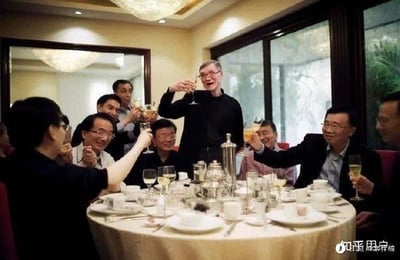 这是一张在中式餐厅包厢内拍摄的纪实照片，画面中比尔·盖茨位于中心位置，与周围多位身着正装的男士一同举杯，桌上摆放着中式餐具与酒具，众人面带笑容，氛围轻松融洽，呈现出一场国际商务交流后的聚餐酒局场景。