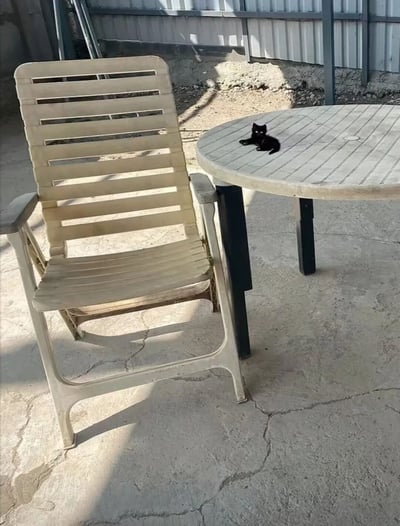 一只通体漆黑的小猫正慵懒地趴在一张圆形木桌上休息，木桌表面有明显的使用痕迹和木纹。旁边放着一把米白色的塑料椅，椅子有竖条状的靠背设计。整个场景在户外的水泥地面上，背景是一面波纹状的金属墙，阳光照射下形成了明显的光影对比。画面传递出一种宁静闲适的氛围。