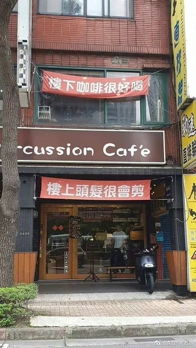 这是一张实拍的街边商铺图，画面中一楼是名为“cussion Caf'e”的咖啡店，店门上方悬挂红色繁体横幅，写着“樓上頭髮很會剪”，为楼上的理发店做宣传；二楼对应的位置悬挂另一张红色繁体横幅，写着“樓下咖啡很好喝”，为楼下的咖啡店做宣传，形成了有趣的店家互助营销场景。咖啡店玻璃门内可以看到一名站立的人员，店门口右侧停着一辆黑色电动车，周围是日常的街道路面与绿植，整体氛围生活化。