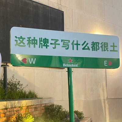 一张户外广告牌的实拍照片，整体采用喜力啤酒标志性的绿白配色方案。牌面上方用绿色大字写着"这种牌子写什么都很土"的自嘲式标语，底部则保留了品牌经典的红色五角星logo和"Heineken"英文标识，左右两侧还有装饰性的红色心形图案和字母"W"、"E"。背景是浅色石材墙面和一块黑色电子屏，营造出一种品牌故意用简陋招牌风格进行自我调侃的幽默效果。