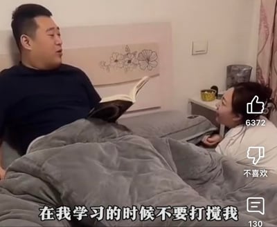 这是一张抖音短视频的截图，画面呈现卧室场景：丈夫靠在床头，盖着灰色被子，手里摊开一本书，对着凑到床边的妻子摆出不耐烦的神情，配文“在我学习的时候不要打搅我”。但从丈夫的状态来看，他根本没有认真看书，只是以此为借口拒绝和妻子互动，形成了搞笑的反差感。画面右侧带有抖音平台的互动界面，显示点赞数6372、评论数130，还有“不喜欢”按钮，进一步说明这是平台上的短视频内容。