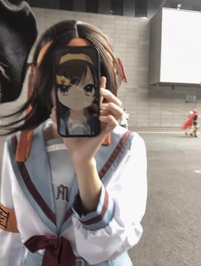 在漫展户外场地中，一位还原《凉宫春日的忧郁》里凉宫春日造型的coser，手持显示着凉宫春日动漫形象的手机，将手机精准对准自己脸部位置，利用视觉错位巧思，让真人cos的发型、服饰与二次元角色的脸部完美衔接，制造出仿佛三次元coser直接“变”成二次元角色的趣味效果，背景还能看到其他漫展参与者的身影，充满ACG爱好者的创意巧思。