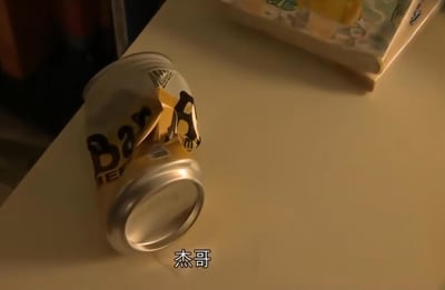 一张桌子上放着一个皱巴巴的Bay Area饮料罐，罐子已经变形，部分标签脱落。罐子旁边有一个模糊的盒子状物体。图片底部有白色文字“杰哥”。