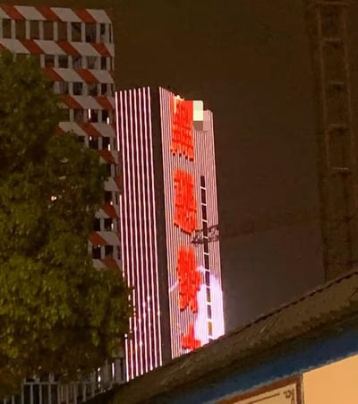 这是一张夜晚拍摄的城市建筑照片，主要展示了一栋高楼大厦。建筑物外墙有红色的大型文字，但部分区域被模糊处理。左侧有一栋带有红白相间条纹的建筑，前景有一些树木。整体画面是夜景，建筑物有灯光照明，营造出城市夜晚的氛围。