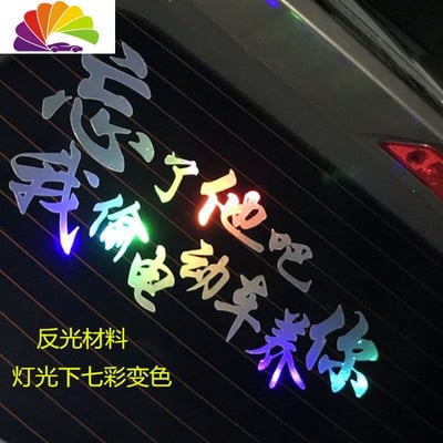 图片展示了一辆汽车的后窗玻璃上贴着一张反光材质的趣味贴纸，上面印有网络流行梗文字“忘了他吧，我偷电动车养你”。文字采用七彩渐变效果，在灯光照射下会呈现变色视觉效果。贴纸下方还有黄色小字标注“反光材料 灯光下七彩变色”，说明其材质特性。该梗源于广西偷车男的经典语录，通过将违法行为幽默化为荒诞承诺，反映了网民面对经济压力时的自嘲与乐观态度。