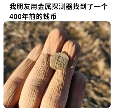 图片上方配有文字“我朋友用金属探测器找到了一个400年前的钱币”，下方是一只手握着一枚表面有模糊纹路的小硬币，背景为模糊的泥土和枯草。硬币外观普通，纹路不清晰，与“400年前”的描述形成反差，营造出幽默调侃的效果，暗示这可能只是一个普通的旧物件而非真正的古董。