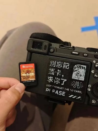这是一张实拍的生活趣味图，画面里一只手捏着一枚任天堂Switch的《塞尔da传说》日版游戏卡带，凑到一台黑色微单相机的机身旁，相机机身贴有醒目的提示贴纸，写着“别忘记插卡，求你了”以及对应的英文提醒，本意是提醒使用者不要忘记带相机的SD存储卡，但图中人物带的却是Switch游戏卡，形成了“带了卡，但带的不是所需的卡”的乌龙效果，完美呼应了标题“带了 但是没带”的笑点。