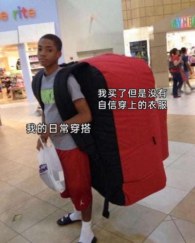 这是一张在商场内拍摄的真人搞笑梗图，图中的黑人少年穿着灰色耐克短袖T恤与红色运动短裤，背着一个尺寸夸张到不合常理的红黑配色背包，手里提着白色购物袋，表情略显窘迫无奈。图片通过两处中文文字标注进行调侃：将少年身上简约普通的日常服饰标注为“我的日常穿搭”，把那个超大号背包标注为“我买了但是没有自信穿上的衣服”，用夸张的比喻手法，生动地映射出当代很多人网购或逛街时买下心仪但风格大胆、超出自己日常风格的衣服后，因不自信而将其闲置的普遍生活状态，内容充满共鸣感与搞笑趣味。